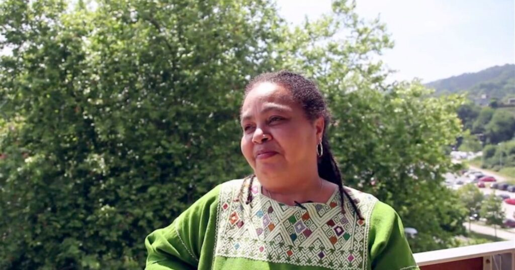Foto mostra Ochy Curiel, mulher negra, de cabelos crespos e presos. Está em ambiente aberto, com folhas de árvores ao fundo. Ela usa um vestido verde com bordados.